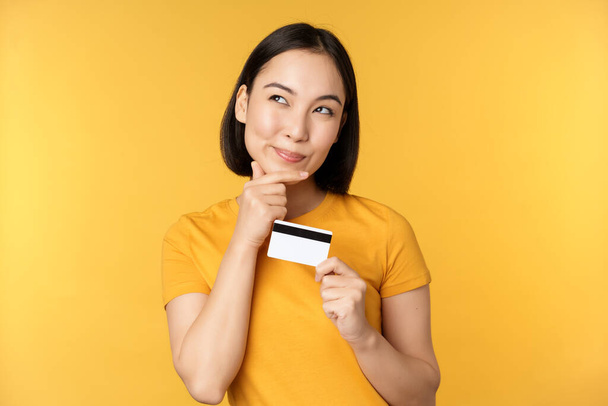Ajatteleva aasialainen nainen, jolla on luottokortti ja hymyilevä, suunnittelee ostaa smth, seisoo huomaavainen yli keltainen tausta - Valokuva, kuva