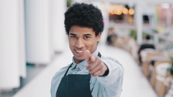 Φιλικός αφροαμερικάνος νεαρός άνδρας σερβιτόρος πωλητής εστιατόριο καφέ κουζίνα μπαρ εργαζόμενος σε ποδιά κοιτάζοντας κάμερα δείχνει χειρονομία της έρχεστε εδώ προσέγγιση ευπρόσδεκτη προσκαλώντας τους πελάτες προσφορές - Πλάνα, βίντεο