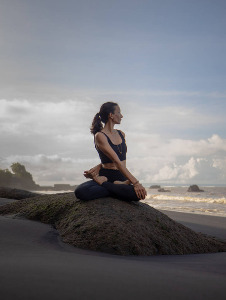 Пляжная йога. Азиатка практикует Parivrtta Sukhasana на открытом воздухе. Сидя в вариации позы Лотоса. Легкая поза. Растягивание бедер, коленей, плеч и позвоночника. Пляж Менгенин, Бали, Индонезия - Фото, изображение