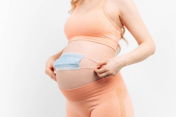Belly of a pregnant woman holding a medical mask close-up on a white background. Pregnancy of a woman and a medical mask on her stomach, the concept of protection from epidemics - Foto, Imagem