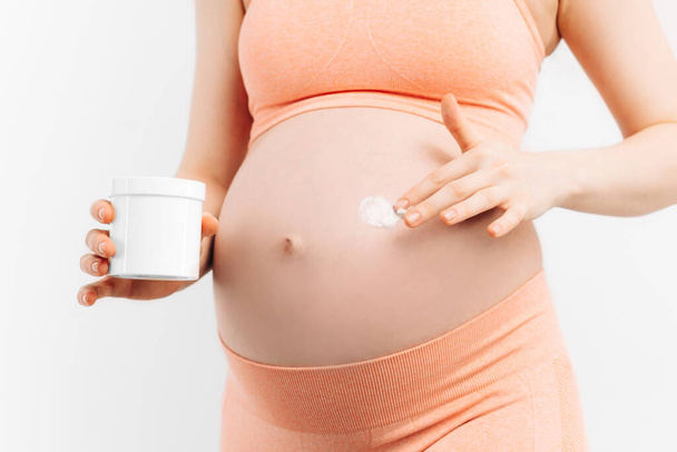 Detailní záběr těhotné ženy aplikující hydratační krém na břicho na bílém pozadí. Koncept těhotenství, mateřství, příprava a očekávání - Fotografie, Obrázek