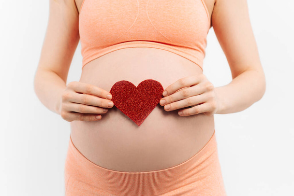 Young pregnant woman holding and touching her belly, holding a red heart sign, belly close-up. birth life concept, happy woman pregnancy, woman holding hands symbol heart white background - Φωτογραφία, εικόνα