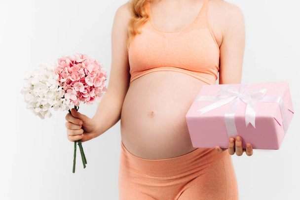 Beautiful pregnant woman with flowers holds her hands on her stomach on a white background. A young woman is waiting for the birth of a child. Pregnancy, motherhood, mother's day holiday concept - Foto, afbeelding