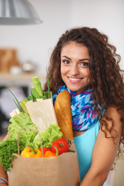 Młoda kobieta trzyma torbę na zakupy z warzywami Stojąc w kuchni. - Zdjęcie, obraz