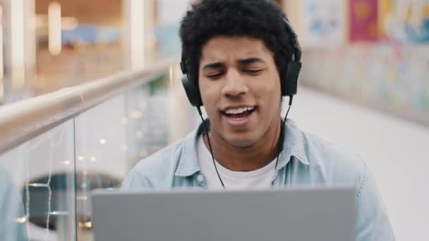 African american 20s guy student business male freelancer sitting in cafe indoors with laptop wears headphones listening to music online website enjoying audio sound in computer app singing song break - Footage, Video