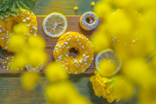Sweet dessert donut covered with lemon glaze, sprinkled with caramel dragee, lemon slices. Spring Easter card. Donuts with sweet yellow glaze with lemon flavor, on texture of board. Flat lay. soft focus - Photo, Image