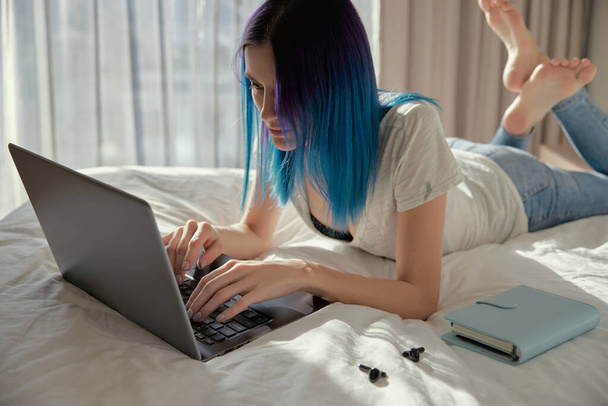 Σοβαρή γυναίκα με μπλε μαλλιά στην κρεβατοκάμαρα εργασίας με φορητό υπολογιστή. - Φωτογραφία, εικόνα