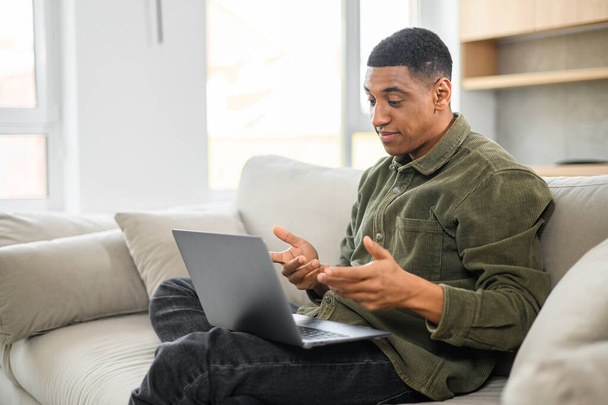 Cheerful multiracial man wearing casual wear sitting on the sofa and using laptop at modern home office. Glad male entrepreneur gesturing while chatting - Φωτογραφία, εικόνα