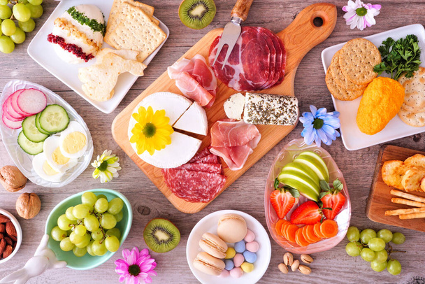 Spring or Easter theme charcuterie table scene against a wood background. Collection of cheese, meat, fruit and vegetable appetizers. Overhead view. - Photo, Image