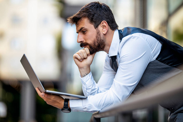 Молодой бизнесмен, работающий онлайн на ноутбуке, за пределами своего офисного здания. - Фото, изображение