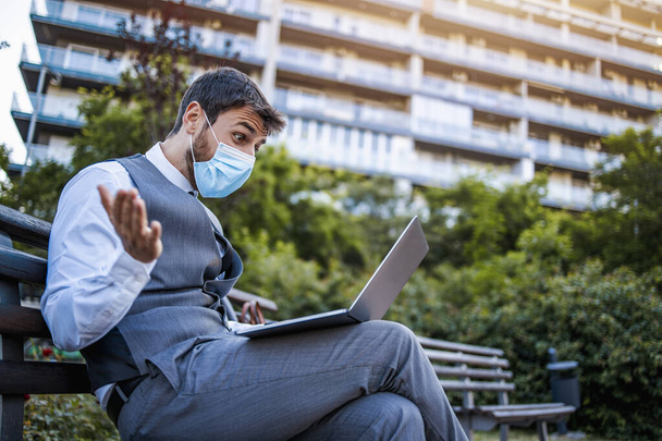 Молодой бизнесмен в защитной маске работает на открытом воздухе над ноутбуком. Защита от эпидемии Covid-19. - Фото, изображение