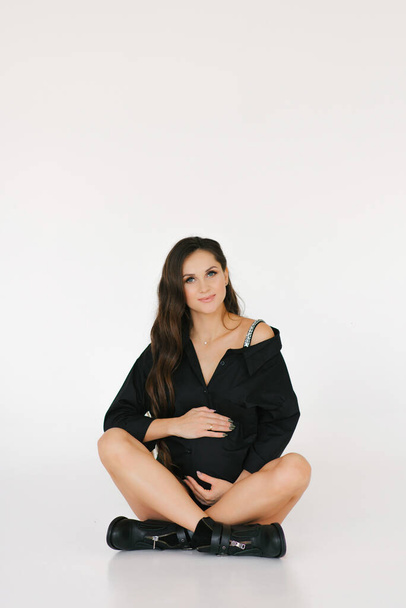 A stylish pregnant woman in a black shirt and boots sits on a white background and hugs her stomach - Photo, Image