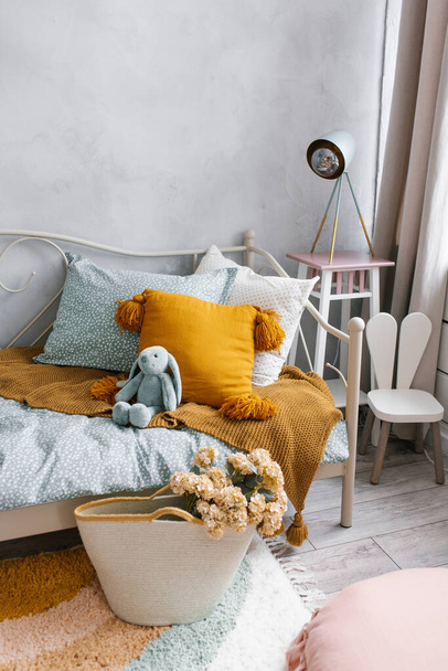 A bed in the children's bedroom with a bunny toy and pillows. Next to the bed is a wicker basket with flowers as a decorative element. Modern interior of the children's room - Foto, Imagen