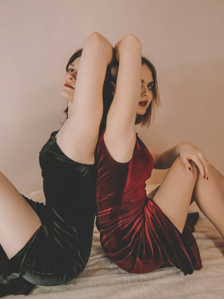 Two attractive slender young women, a brunette and a brown-haired woman in evening velvet dresses of emerald and burgundy colors, with bright evening makeup and bright red manicure, sit back to back on a beige blanket against a light wall - Fotografie, Obrázek