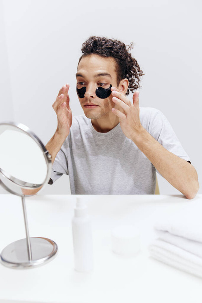 tipo con el pelo rizado en una camiseta blanca se sienta en una mesa con un espejo y una toalla parches para la cara cuidado de la piel - Foto, Imagen