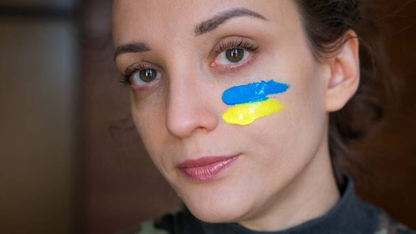 Retrato interior de niña con bandera ucraniana azul y amarilla en la mejilla con uniforme militar, reclutamiento obligatorio en Ucrania, conceptos de igualdad - Foto, Imagen