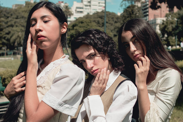 τρία όμορφα νεαρά κορίτσια από την Ισπανία, Λατίνα Αργεντίνισσες, που στέκονται σε ένα πάρκο και ποζάρουν αγγίζοντας τα πρόσωπά τους, ντυμένες με ρετρό ρούχα, vintage και γυναικεία ιδέα.. - Φωτογραφία, εικόνα