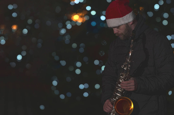 Człowiek grający na saksofonie przed choinką na placu. Facet ma na sobie świąteczny kapelusz. Jest wieczór i wszędzie jest ciemno. Zapalane są tylko lampki i dekoracje świąteczne. - Zdjęcie, obraz