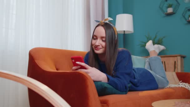 Vidám fiatal felnőtt nő fekszik a kanapén okostelefon videó nézés, online vásárlás, érintés - Felvétel, videó