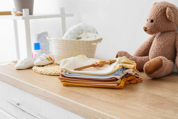 Stack of baby clothes and rattle on wooden chest of drawers in children's room - Photo, Image