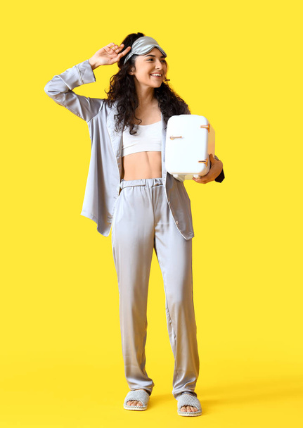 Young smiling woman in pajamas and sleep mask holding cosmetic refrigerator on yellow background - Photo, Image