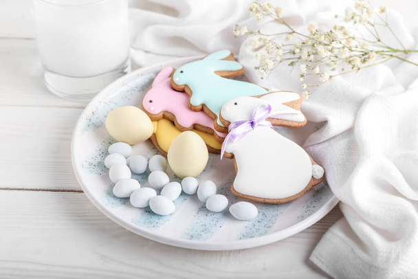 Plate with tasty Easter cookies in shape on bunny, candies and gypsophila flowers on white wooden background - Фото, изображение