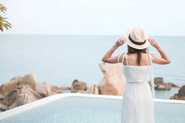 白いドレスと帽子の幸せな旅行者の女性は、若い女性がスイミングプールに立って美しい海の景色をお楽しみください。自由、リラックス、休暇、夏の旅行のコンセプト - 写真・画像