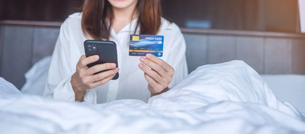 nainen, joka käyttää älypuhelinta ja luottokorttia verkkokauppaan tehdessään tilauksen sängyssä aamulla kotona. teknologia, verkkokauppa, digitaalinen pankkitoiminta online-maksu ja asunto elävä käsite - Valokuva, kuva