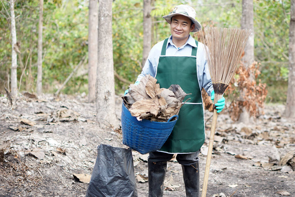 Asian man is holding  broom, basket,garbage bag to get rid of dry leaves in the forest before the summer bushfires. Concept : Forest fire prevention. environmental protection.   - Фото, изображение