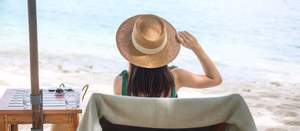 Happy traveler woman in green swimwear and hat enjoy beautiful sea view, young woman sitting on chair at sand beach. Freedom, relaxing, vacation holiday and summer travel concept - Photo, Image