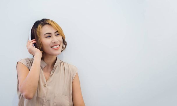 Mulher asiática atraente com cabelo curto em camisa bege sem mangas gosta de ouvir música, podcast ou música no rádio com fone de ouvido branco com sorriso feliz no fundo branco com espaço de cópia. - Foto, Imagem