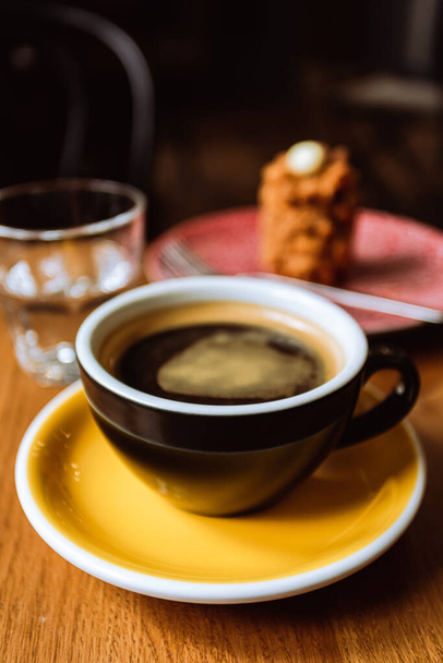 A ceramic cup of black coffee,a dessert served on a plate and a glass of water on the wooden table in a cafe, close-up vertical image - Fotoğraf, Görsel