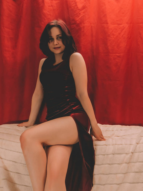 Beautiful young brunette woman with bright makeup with a beautiful slender figure in a velvet burgundy evening dress with a low cut at the hip posing on the back of a sofa in the bedroom, boudoir photo, style and fashion, - Φωτογραφία, εικόνα