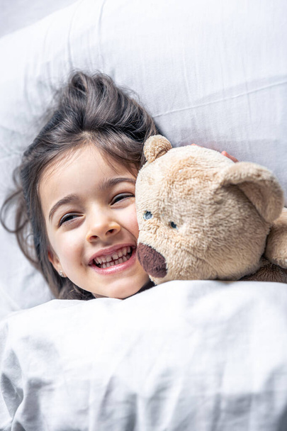 Klein schattig meisje in bed met een teddybeer vroeg in de ochtend. - Foto, afbeelding