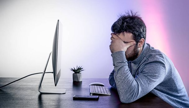 Ένας κουρασμένος άνθρωπος κάθεται μπροστά από έναν υπολογιστή, καλύπτοντας το πρόσωπό του με τα χέρια του. - Φωτογραφία, εικόνα