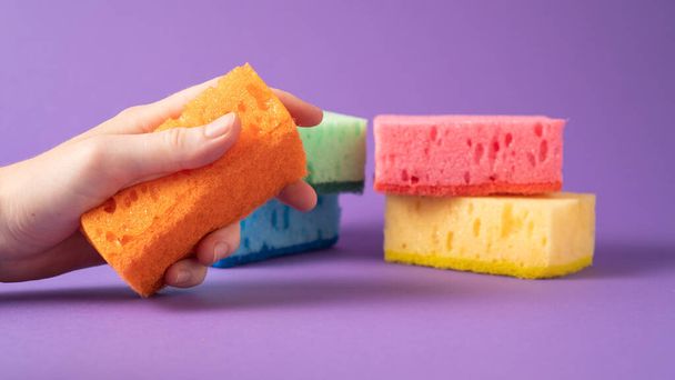 Hand met oranje spons op paarse achtergrond. Klaar voor de afwas. Keukenreinigingssponzen - Foto, afbeelding