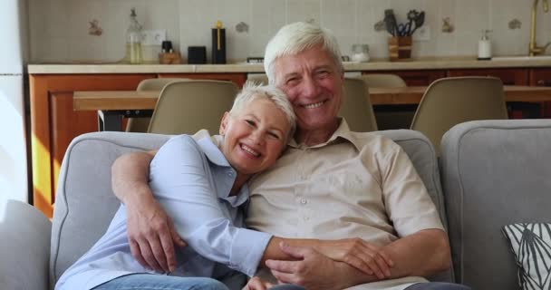 Laughing older couple hugging relaxing on sofa look at camera - Materiaali, video