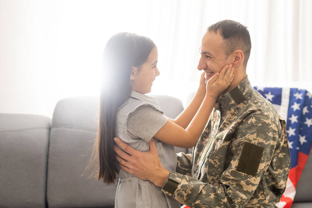 Portrait de heureux père de famille américain en uniforme militaire et jolie petite fille fille avec drapeau des États-Unis étreignant et souriant à la caméra - Photo, image