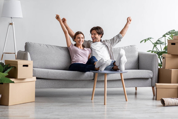 Satisfied caucasian millennial couple sit on sofa, rest, raise their hands up and celebrate purchase of new apartment - Foto, Imagen