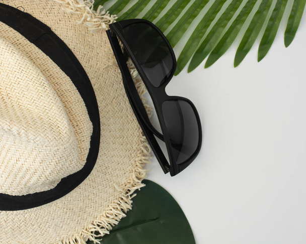 літній світловий капелюх з чорною стрічкою та темними окулярами на білому тлі з пальмовим листям, концепцією морського відпочинку та туризму. плоский простір і копіювання
. - Фото, зображення