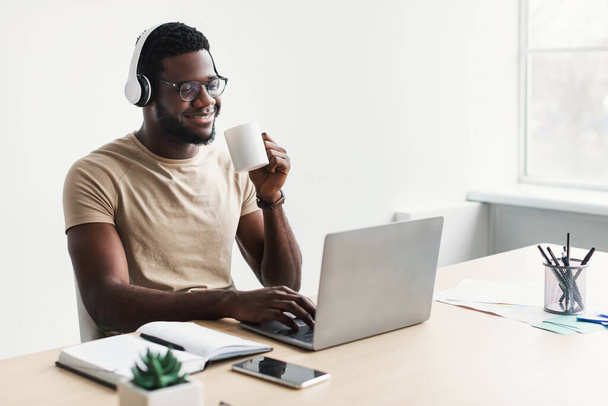 Θετικός νεαρός μαύρος με ακουστικά πίνοντας καφέ, χρησιμοποιώντας φορητό υπολογιστή στο γραφείο στο σπίτι, αντιγραφή χώρου - Φωτογραφία, εικόνα