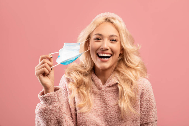 Coronavirus is over. Happy young caucasian lady taking off her face mask, standing on pink studio background - Photo, Image
