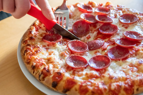 Chlapec ruce krájet salám pizza na kousky s vidličkou a nůž na pizza talíř v close-up makro pohled s lahodným sýrem a salámem jako tuk nezdravé rychlé občerstvení pro hladové teenagery italská pochoutka strava - Fotografie, Obrázek