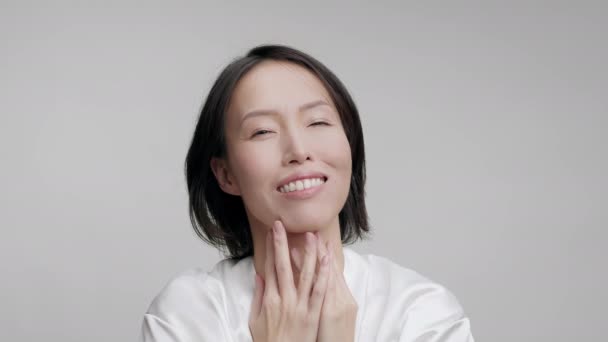 Feliz mujer asiática tocando la cara acariciando mejillas sobre fondo gris - Imágenes, Vídeo