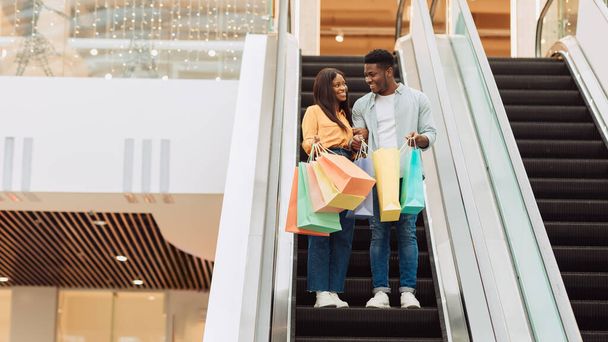 Happy black couple holding shopping bags going on escalator - 写真・画像