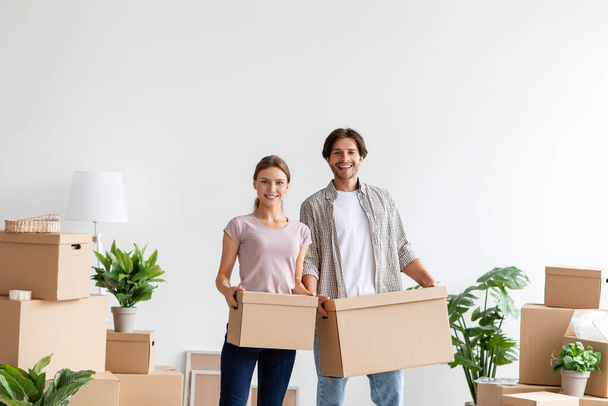 Satisfied european millennial lady and guy in casual carry cardboard boxes with belongings in room, empty space - Photo, Image