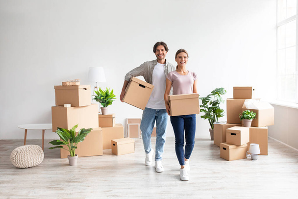 Szczęśliwy młody Europejczyk para w casual nosić kartonowe pudełka z rzeczy w pokoju i cieszyć się zakupem nowego mieszkania - Zdjęcie, obraz