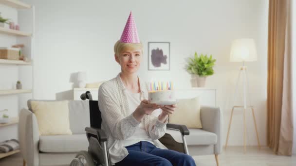 Femme mature positive avec handicap portant un chapeau de fête soufflant des bougies sur le gâteau d'anniversaire et souriant à la caméra, zoom avant - Séquence, vidéo