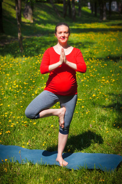 Ejercicio de yoga en el embarazo - mujer embarazada haciendo asana vrikshasana árbol posan al aire libre en césped de hierba con dientes de león en verano - Foto, imagen