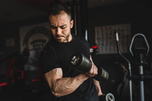 One man young adult caucasian male bodybuilder training arms bicep flexing muscles with dumbbell while standing in the gym wearing black shirt dark photo real people copy space front view waist up - Photo, Image